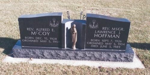 A black granite double gravestone in a grassy cemetery, featuring the names of two priests and a central statue.