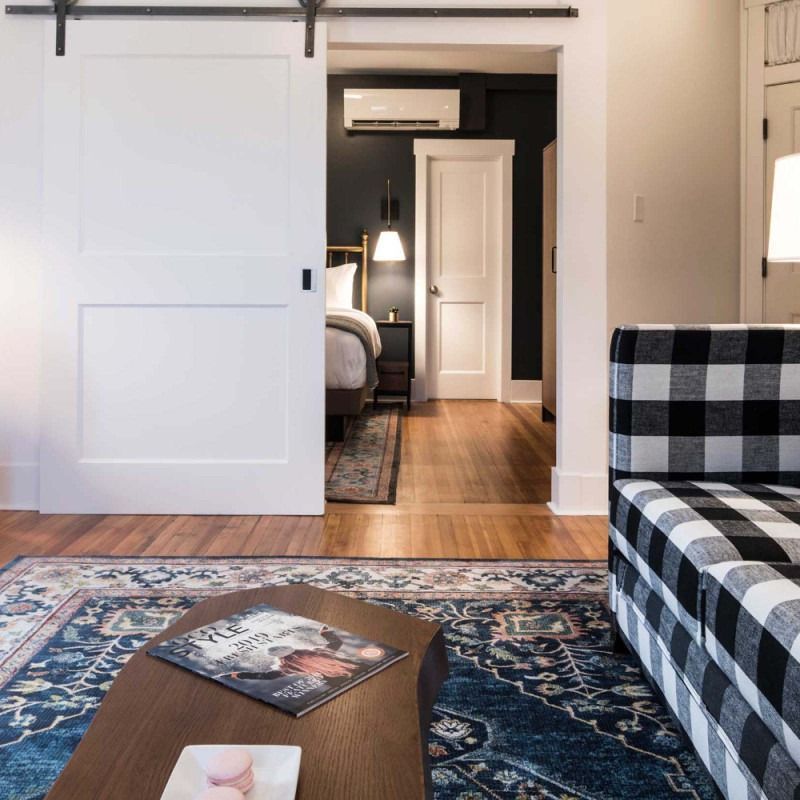 A cozy room with a buffalo check sofa, a wooden coffee table on a blue patterned rug, and a sliding door leading to a bed.