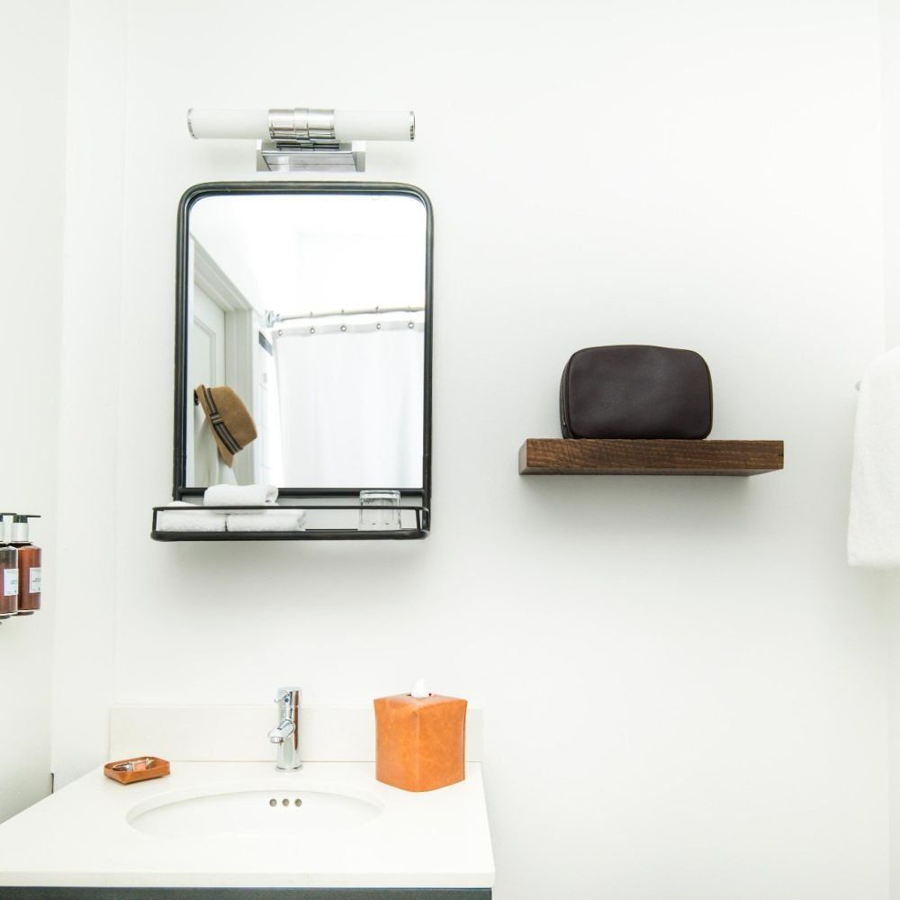 A modern bathroom with a rectangular mirror, a wooden shelf with a pouch, a white sink, and a tan tissue box.