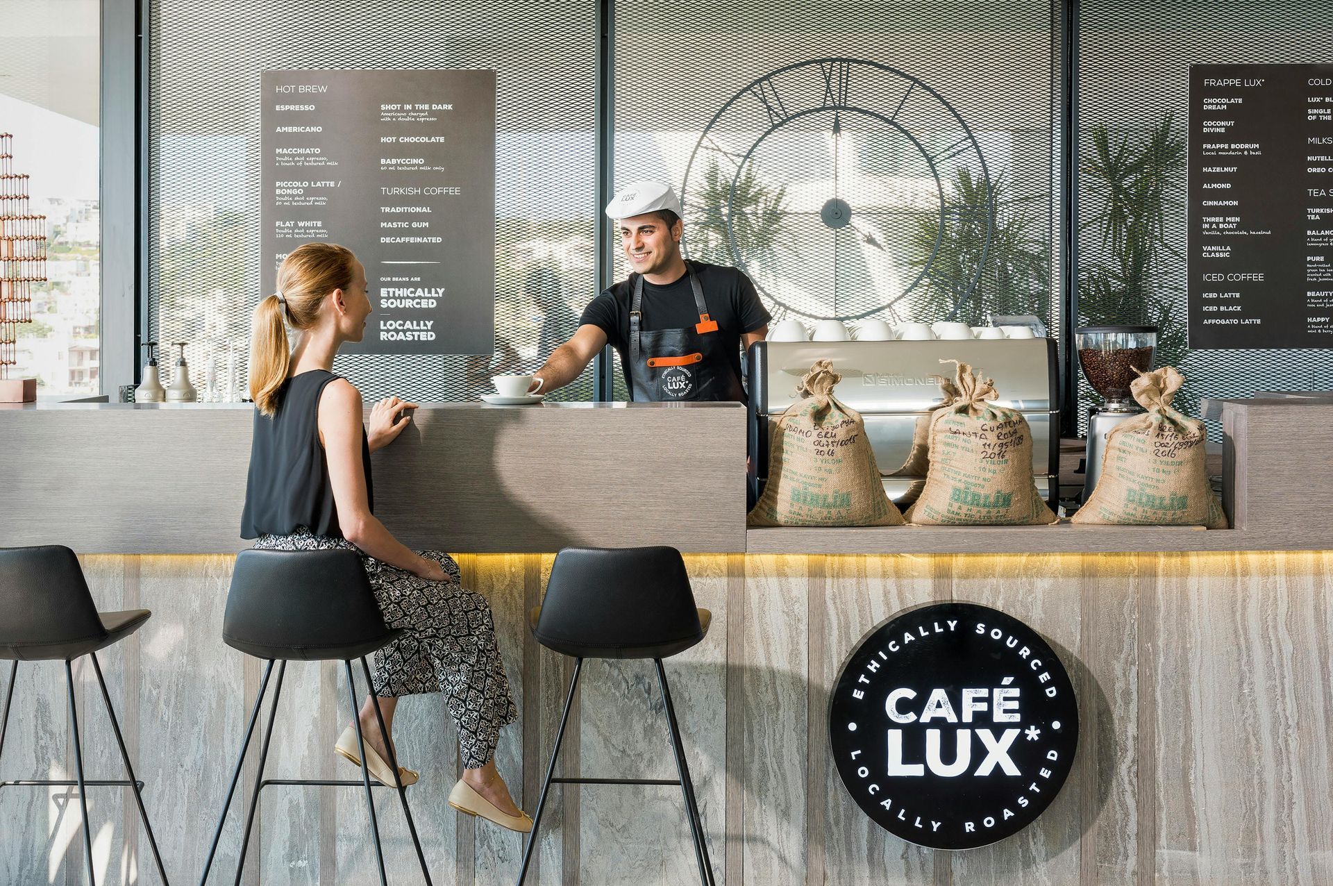 A barista serves coffee to a customer sitting at a Café Lux counter in a modern, brightly lit cafe.