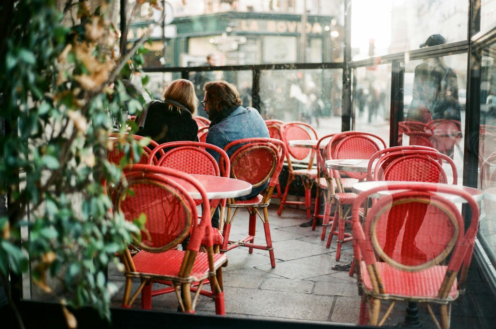 Two people sit at a small table at an outdoor Parisian cafe with red woven chairs and view of a street.