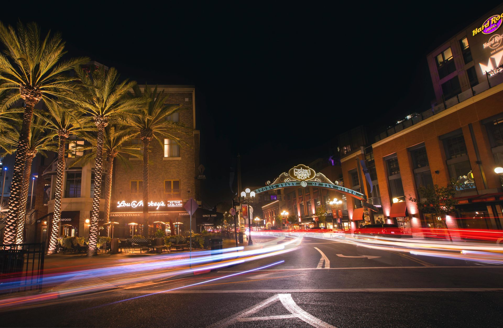 A long-exposure night shot of a San Diego Gaslamp Quarter street, featuring light trails, palm trees, and an arched sign.