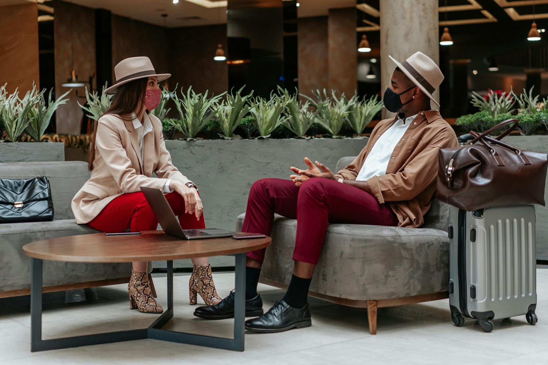 Two people wearing masks and fedoras sit on chairs in a lounge with a laptop, luggage, and plants in the background.