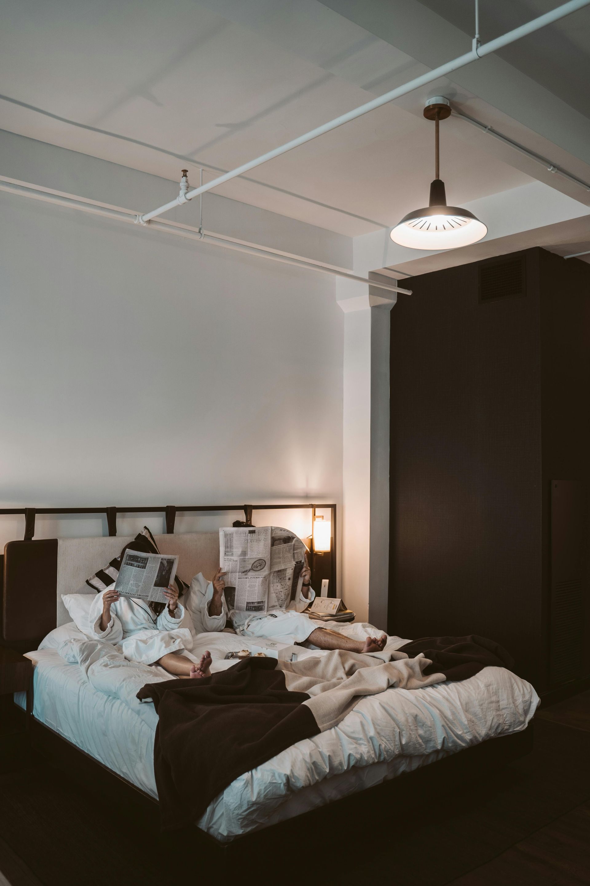 Two people in white bathrobes sit in a bed, reading newspapers in a modern room with a dark accent wall and pendant light.