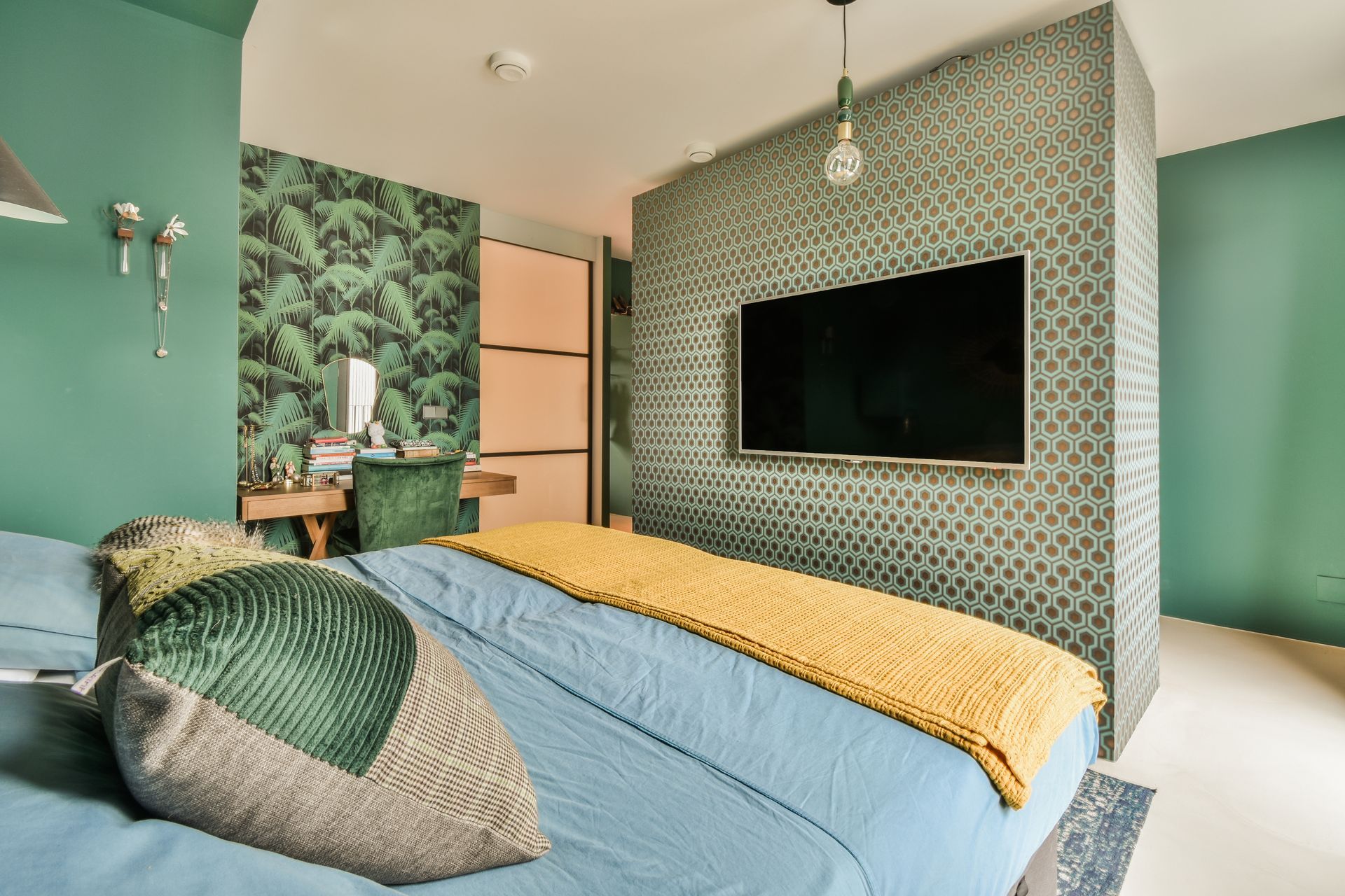 A bedroom with a green wall, patterned wallpaper, a television mounted on a textured wall, and a bed with a yellow throw.
