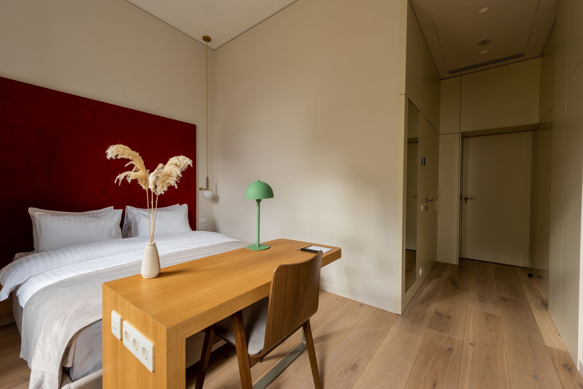 A minimalist hotel room with a bed against a red wall, a wooden desk with a green lamp, and light wood flooring.