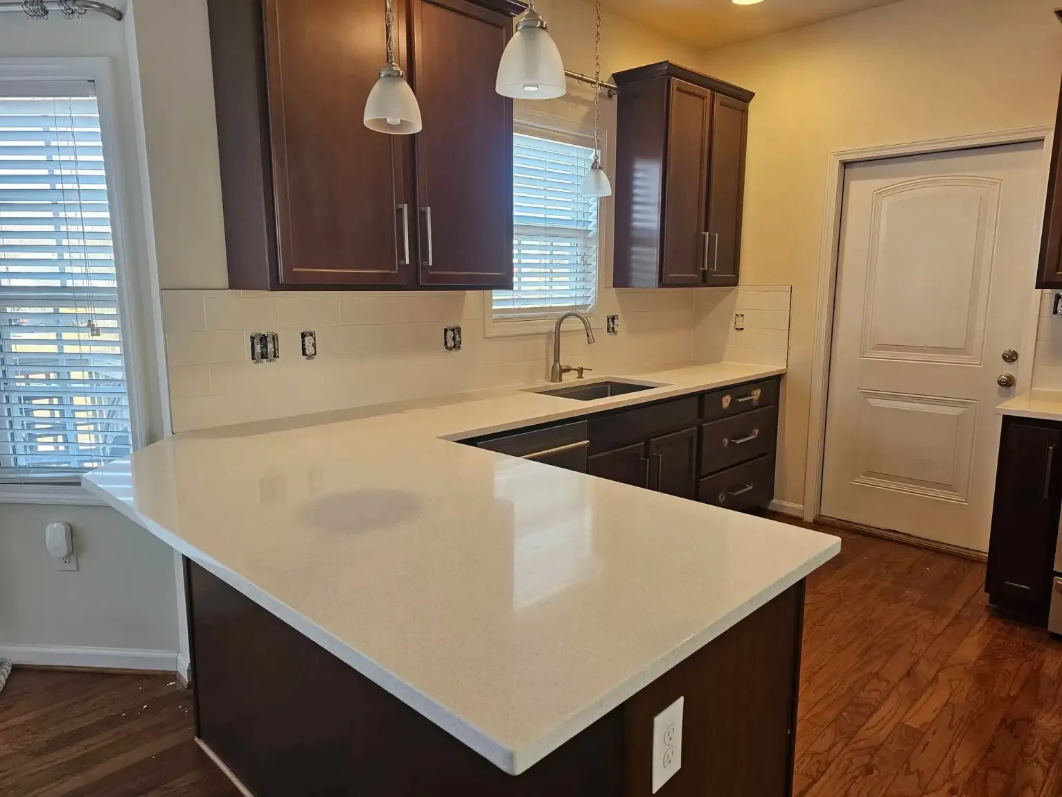 Empty kitchen with dark wood cabinets, white counters, pendant lights, and hardwood floor.