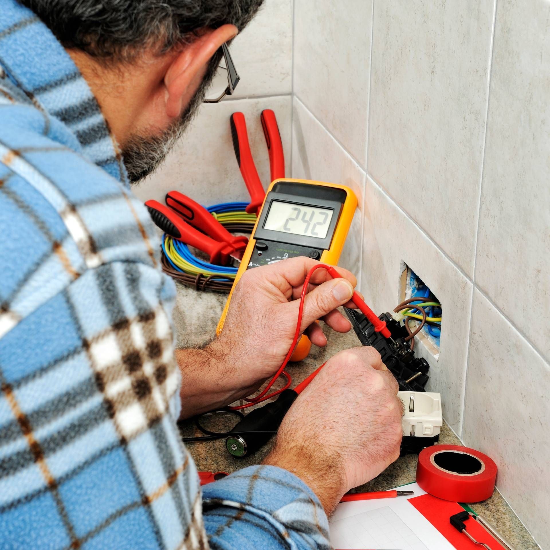 Electrician using a multimeter on an outlet; tools and wires are nearby.