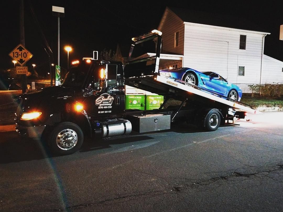 Black tow truck at night with a blue sports car on its bed, beside a building.