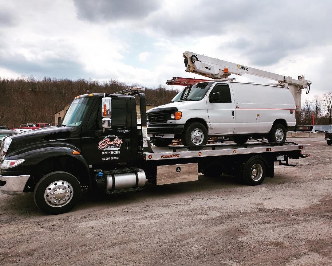 Tow truck with a white van on its flatbed, parked on a gravel lot under a cloudy sky.