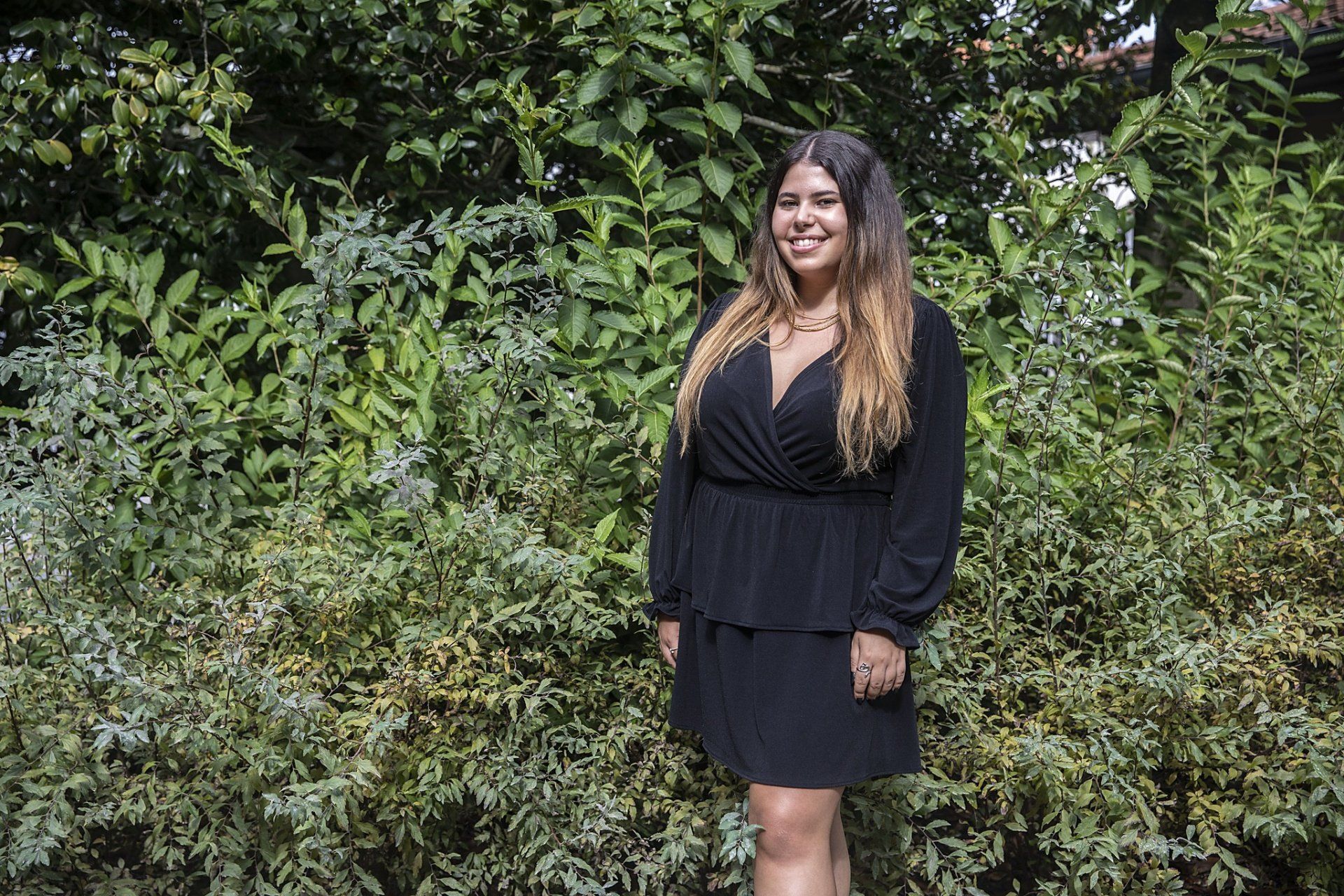 Uma mulher de vestido preto está parada em frente a um arbusto.