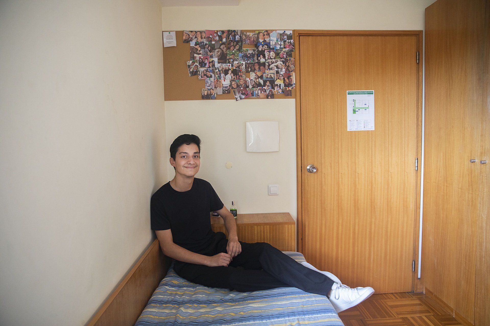 Um jovem está sentado em uma cama em um pequeno quarto.