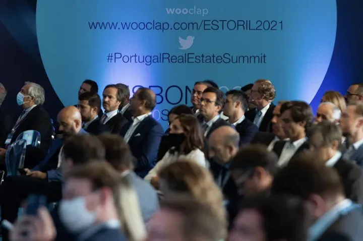 Um grupo de pessoas está sentado em frente a uma placa que diz portugal real estate summit