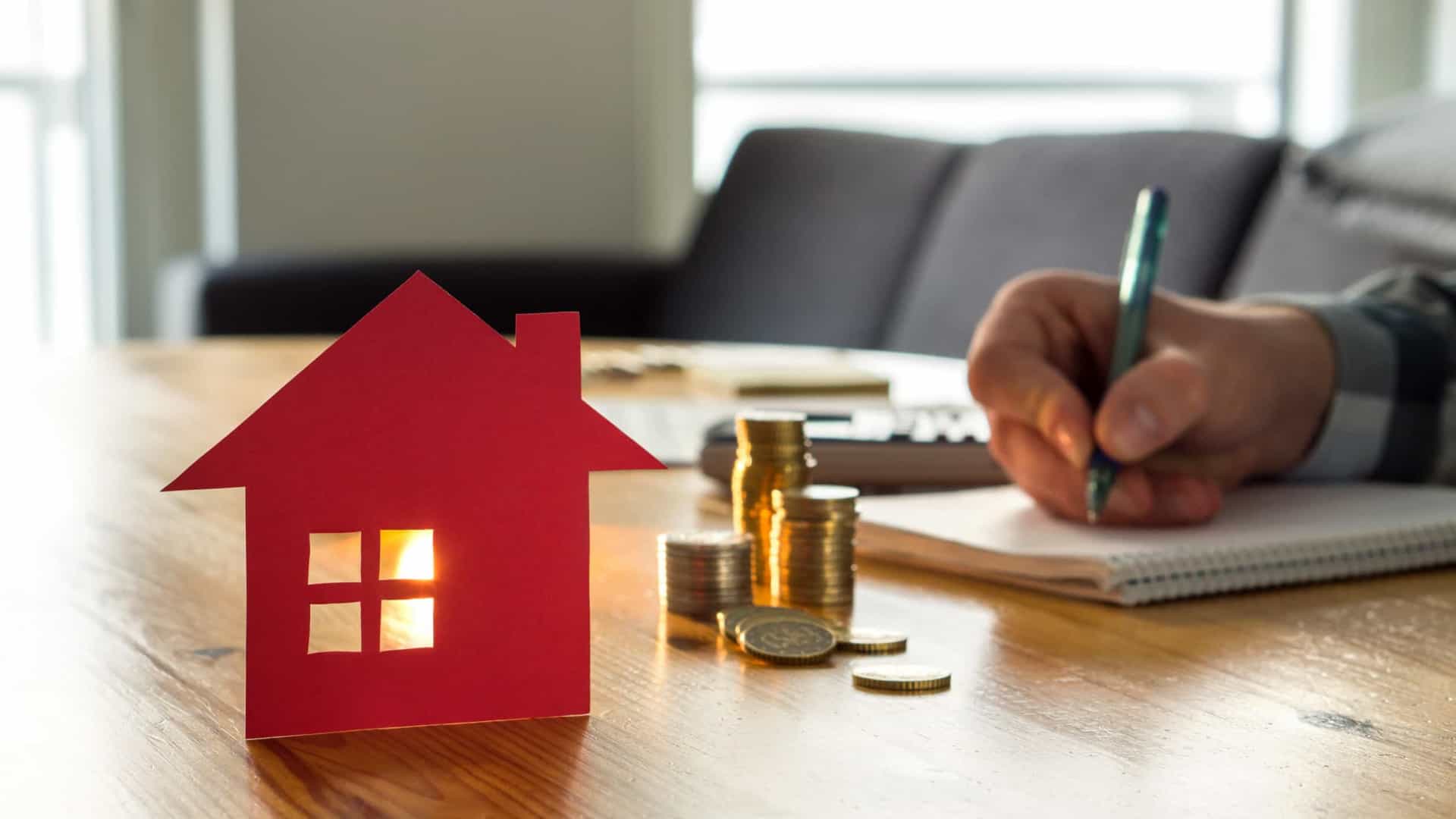 Uma pessoa está escrevendo em um caderno ao lado de uma casa modelo e pilhas de moedas.