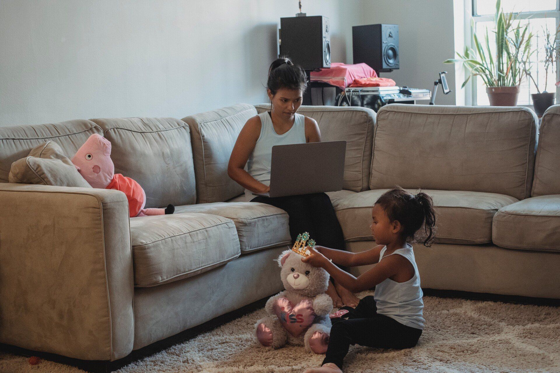 Uma mulher está sentada em um sofá usando um laptop enquanto uma menina brinca com um ursinho de pelúcia.