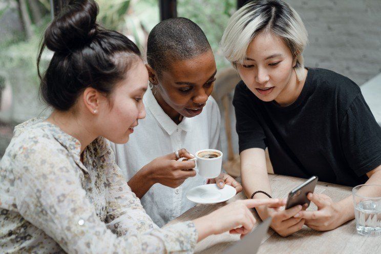 Três mulheres estão sentadas à mesa olhando para um celular.