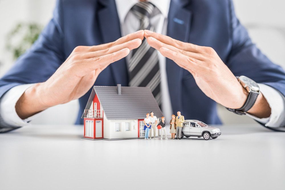 Um homem de terno e gravata está cobrindo uma casa e um carro com as mãos.