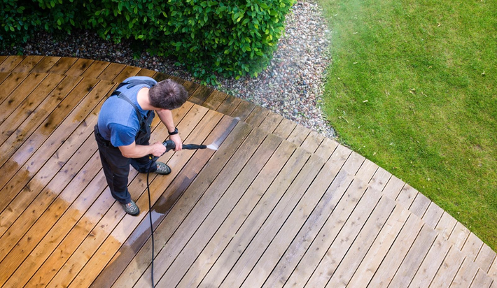 Person power-washing a wooden deck beside a green lawn and hedge