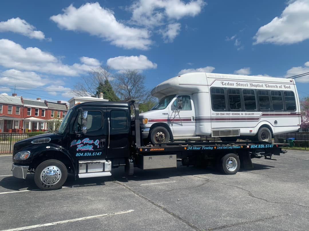 A black tow truck with a white bus on its flatbed, parked in a lot under a blue sky.
