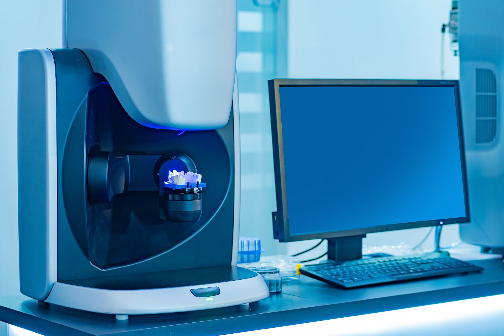 A dental scanning machine with a blue lit interior next to a computer monitor in a lab. — AB Esthetic Dental Laboratory in Robina, QLD