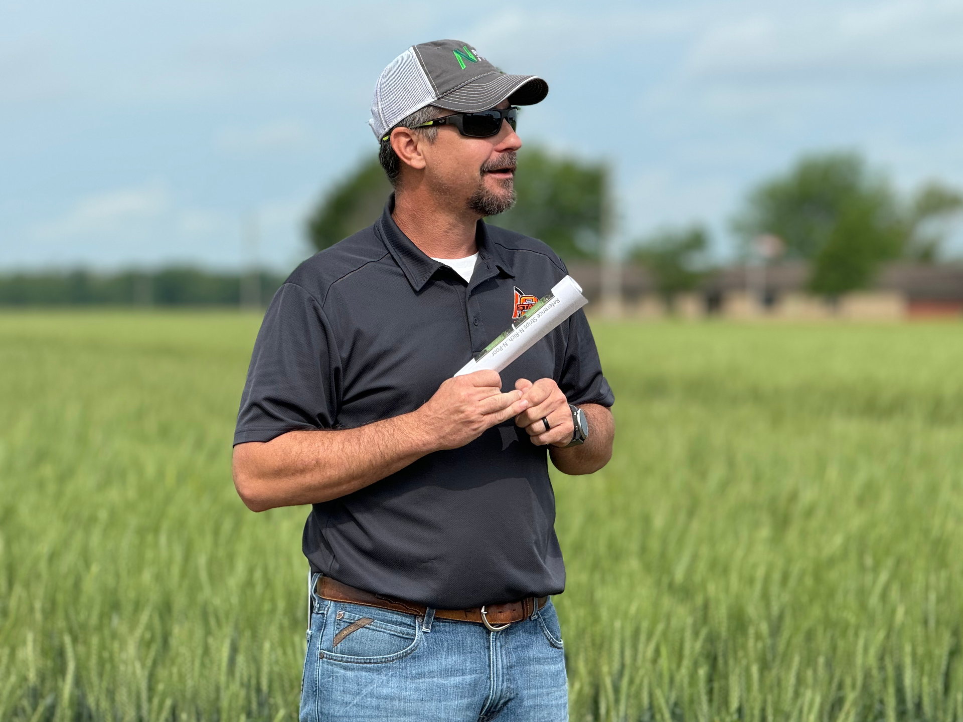 Man standing in a green field holding a white object, wearing a black shirt, cap, and sunglasses.