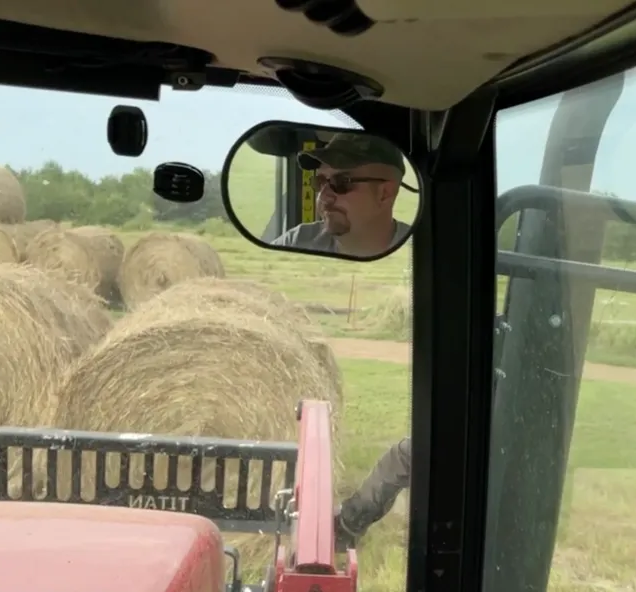 Inside a tractor cab, a man looks out over hay bales in a field.