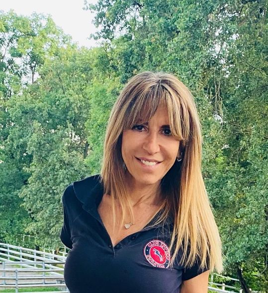 Donna sorridente con una camicia nera in piedi all'aperto con alberi sullo sfondo.