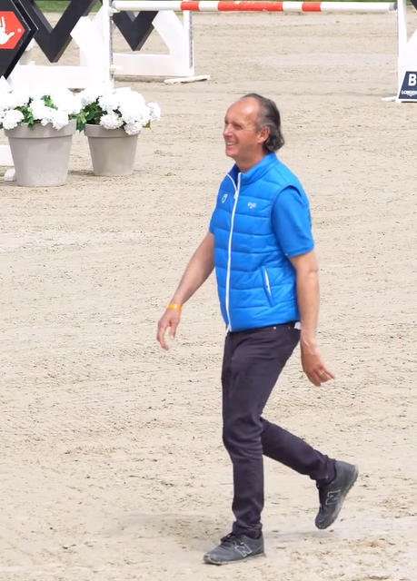 Un uomo con un gilet blu cammina su un'arena sabbiosa vicino a vasi di fiori bianchi.