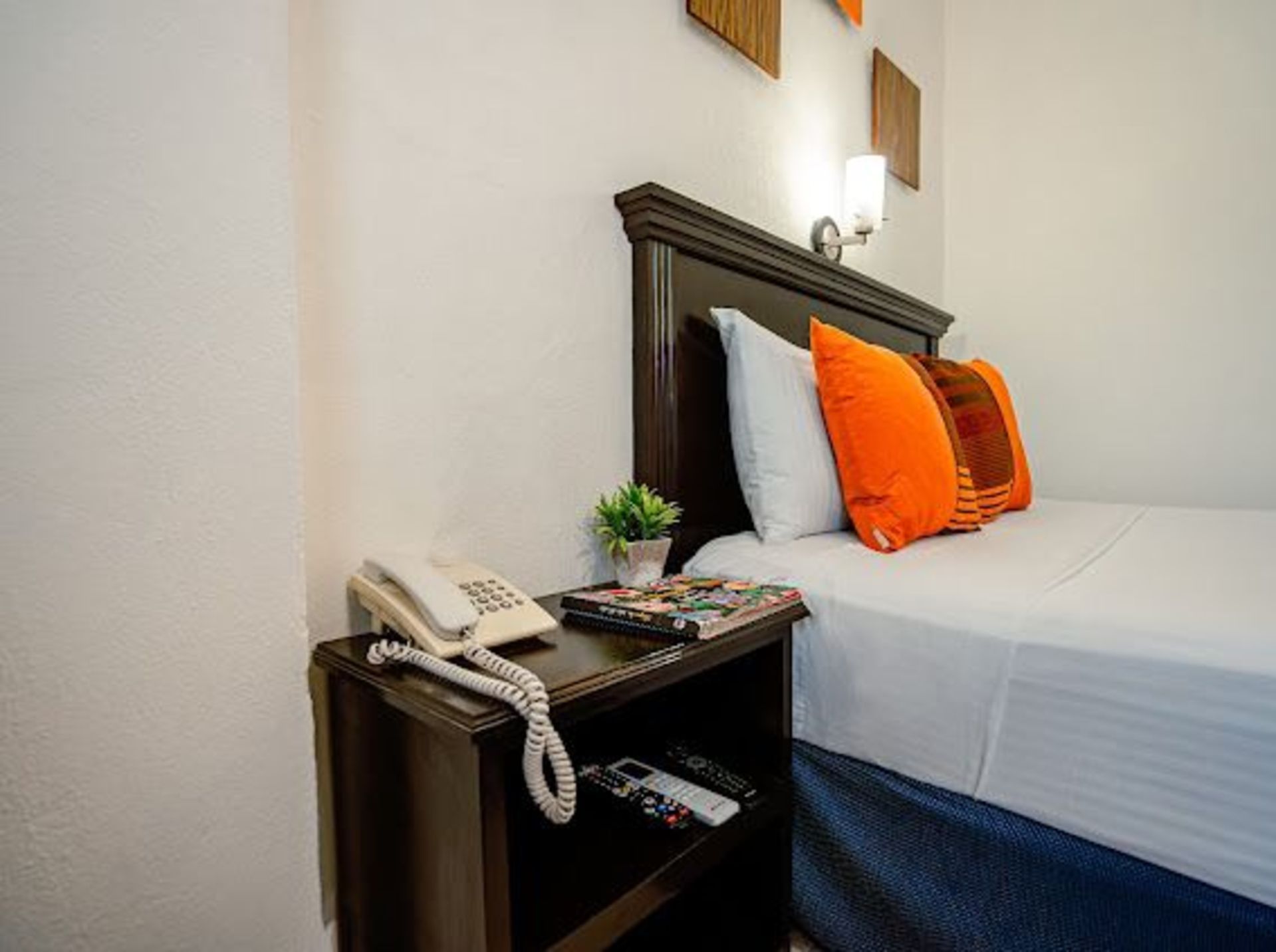Mesita de noche con teléfono blanco, pequeña planta y revistas junto a una cama con almohadas.