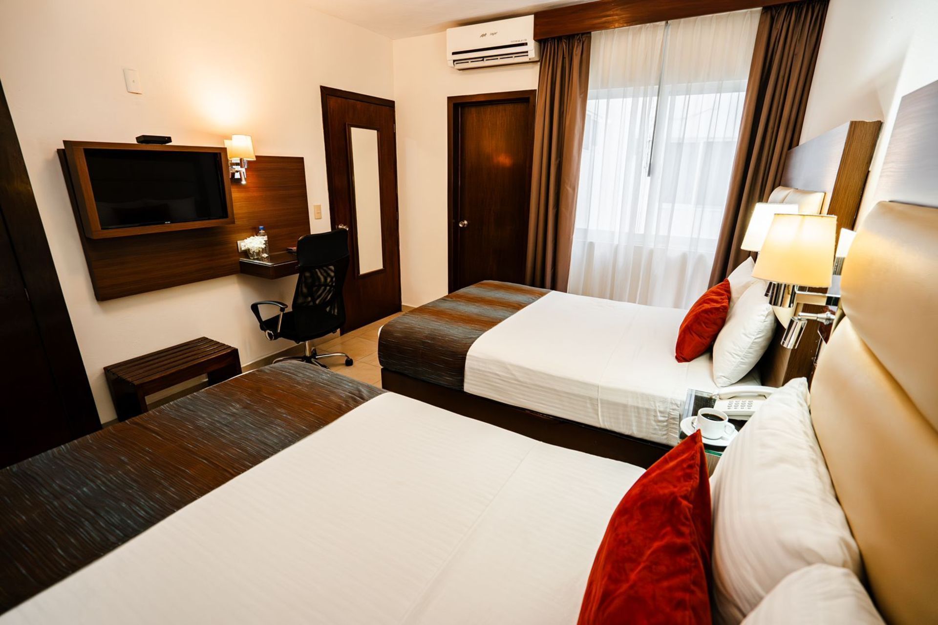 Habitación de hotel con dos camas, almohadas rojas, escritorio, TV y aire acondicionado.