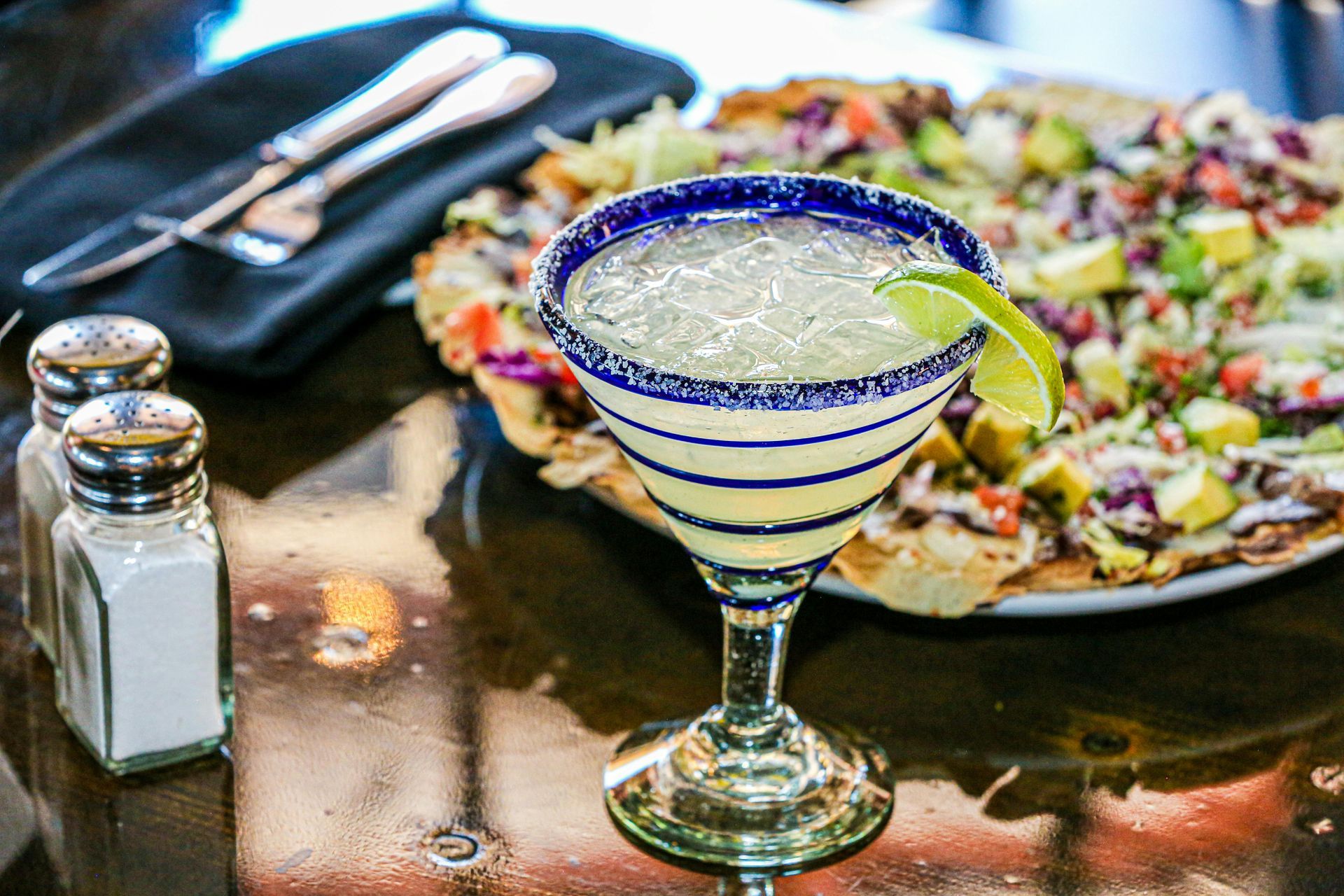 Margarita en vaso con borde azul, gajo de lima, nachos de fondo, salero y pimentero en la mesa.