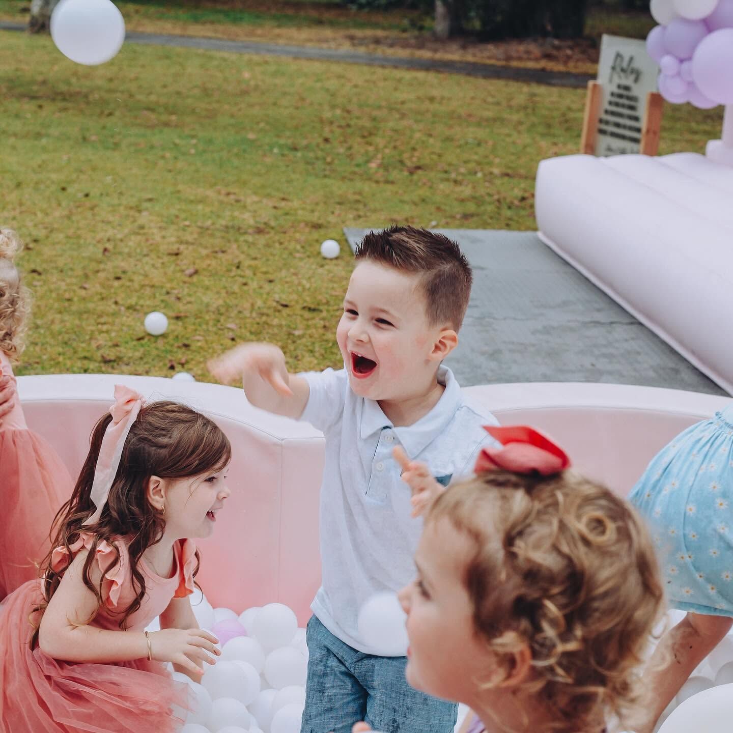 Children playing at a pastel outdoor party, with balloons and a white inflatable backdrop.