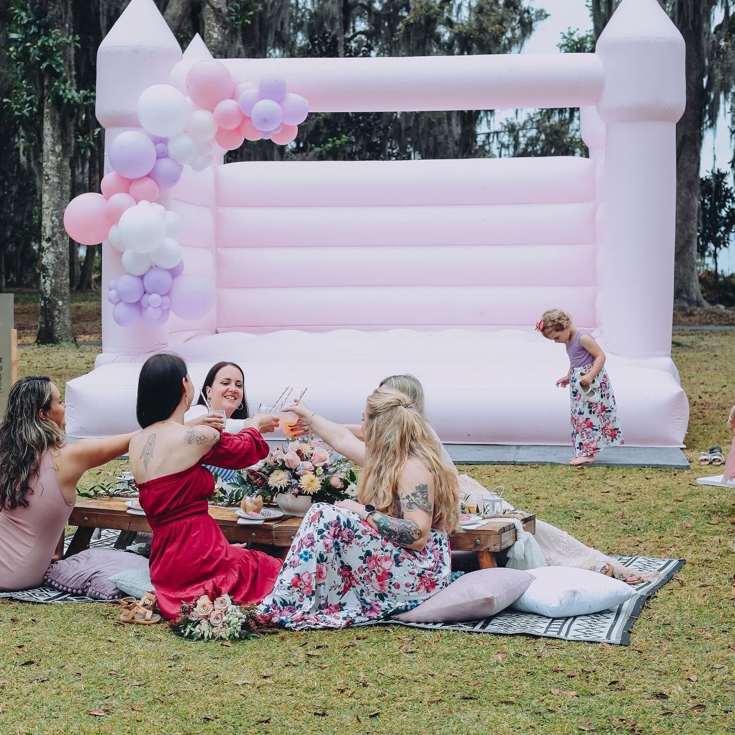 Women sitting on a picnic blanket beside a pink inflatable castle and balloon arch in a park