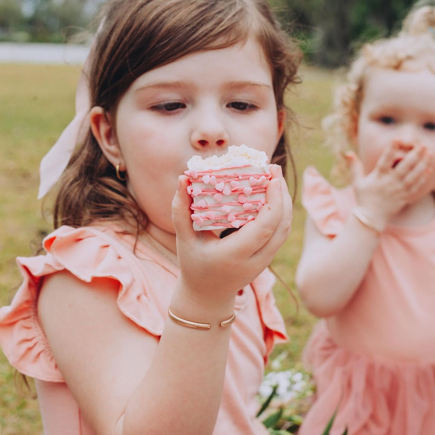 Two children in pink dresses, one eating a frosted cupcake outdoors.