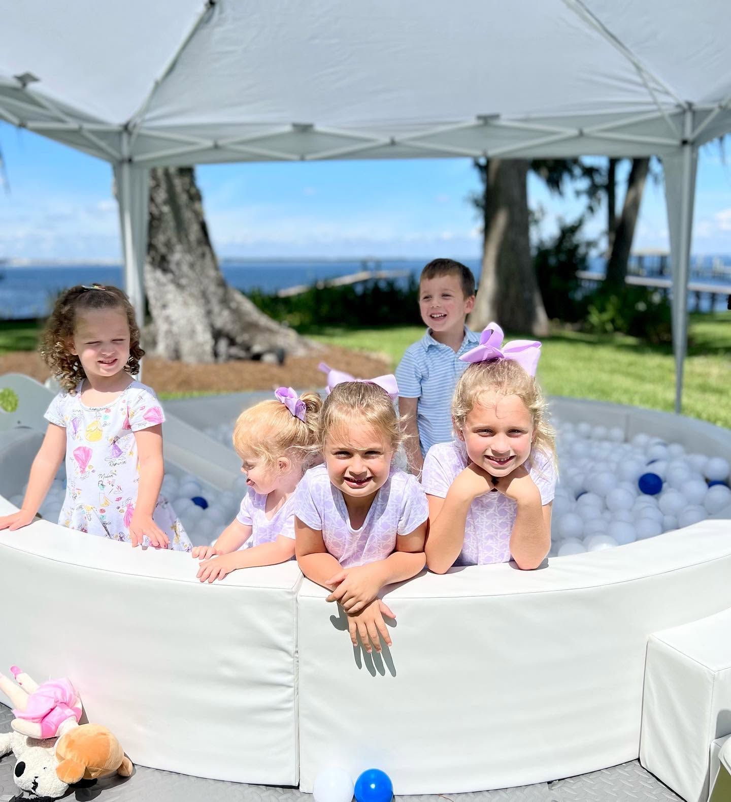 Five children smiling in a white inflatable pool under a canopy outdoors