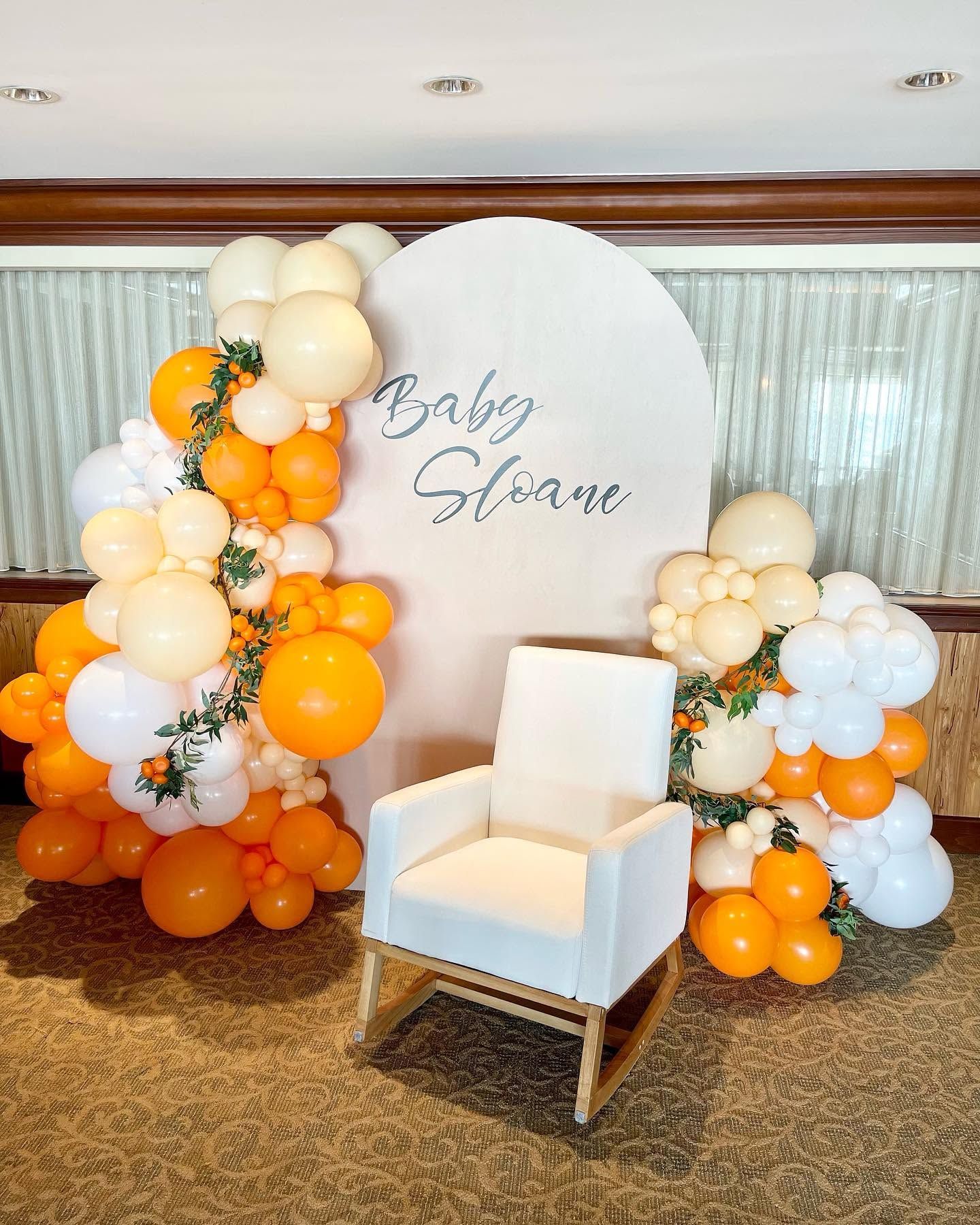Baby shower backdrop with white, orange, and cream balloons and a white chair in front