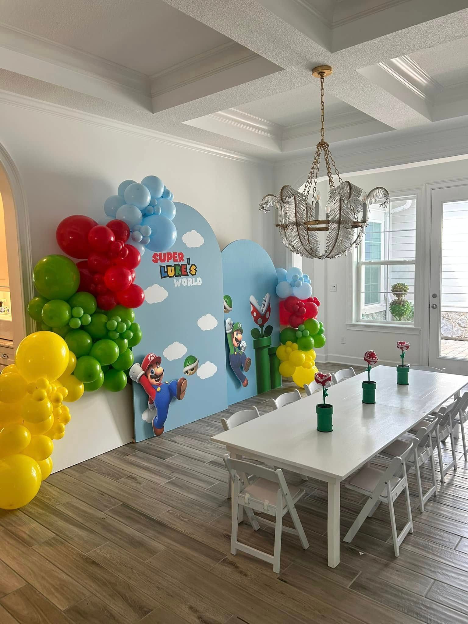 Balloon-decorated party room with a long white table, chandelier, and blue backdrop by a window.
