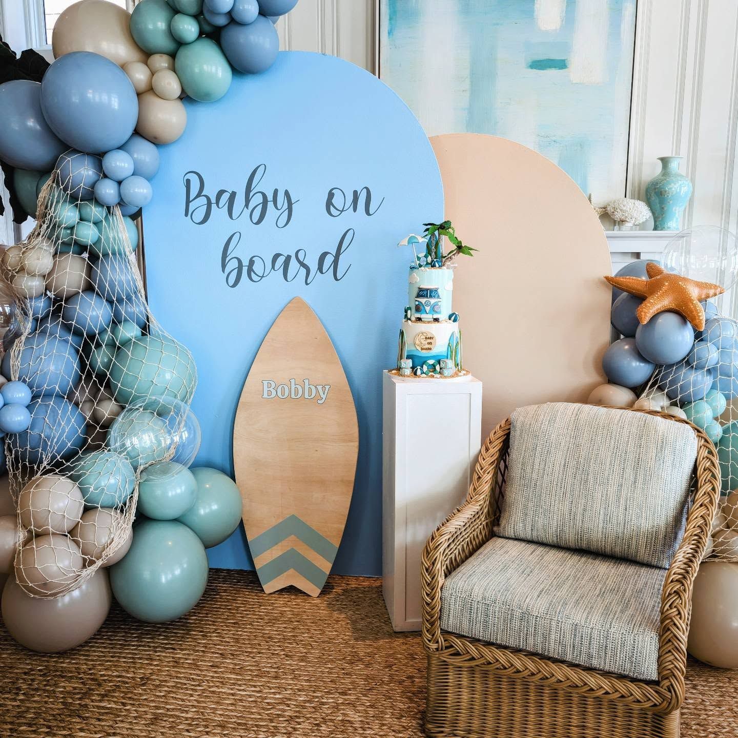 Baby shower display with blue balloons, surfboard sign, cake, and chair on a woven rug