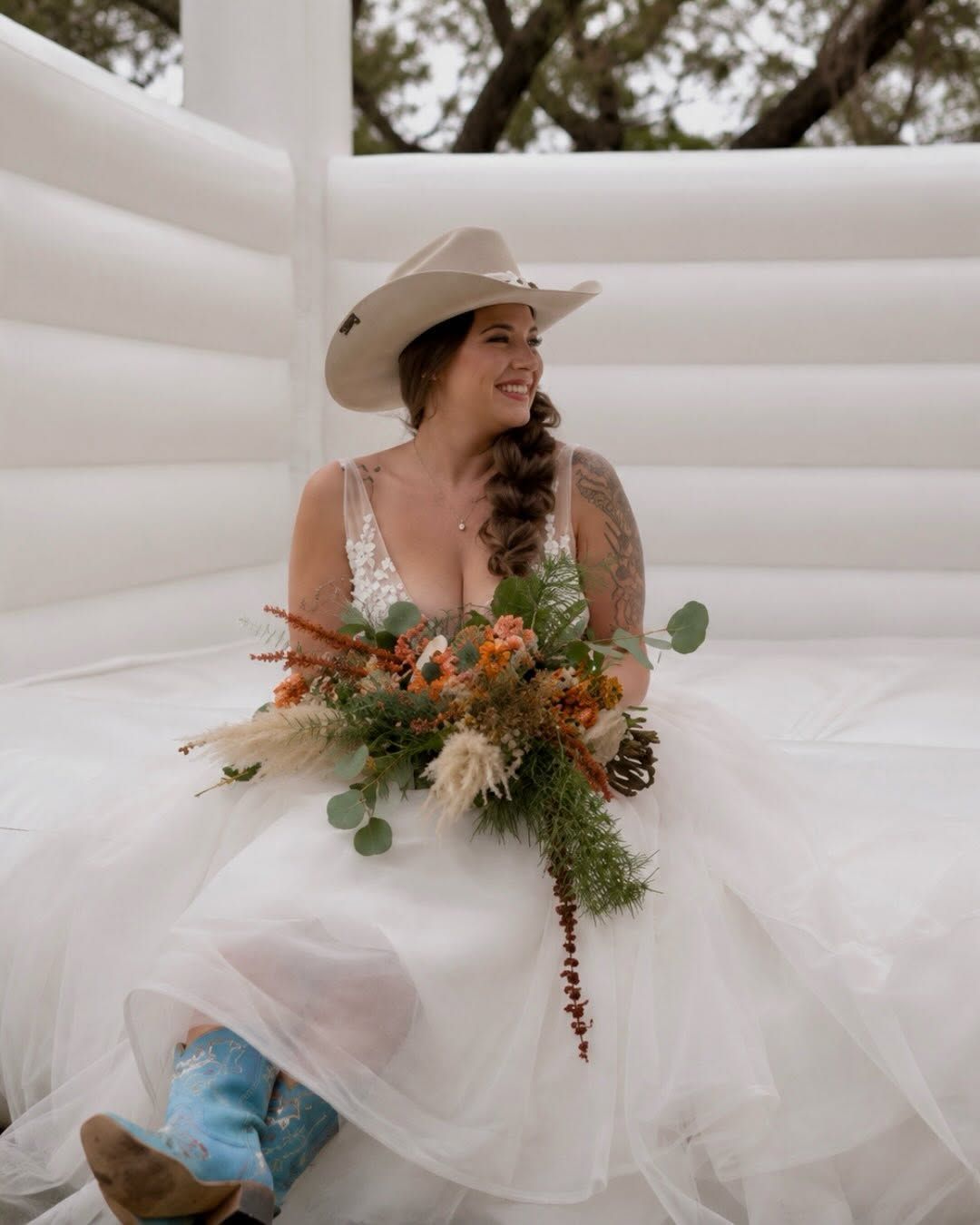 Woman in a white cowboy hat and dress sits with a bouquet inside a white inflatable bounce house
