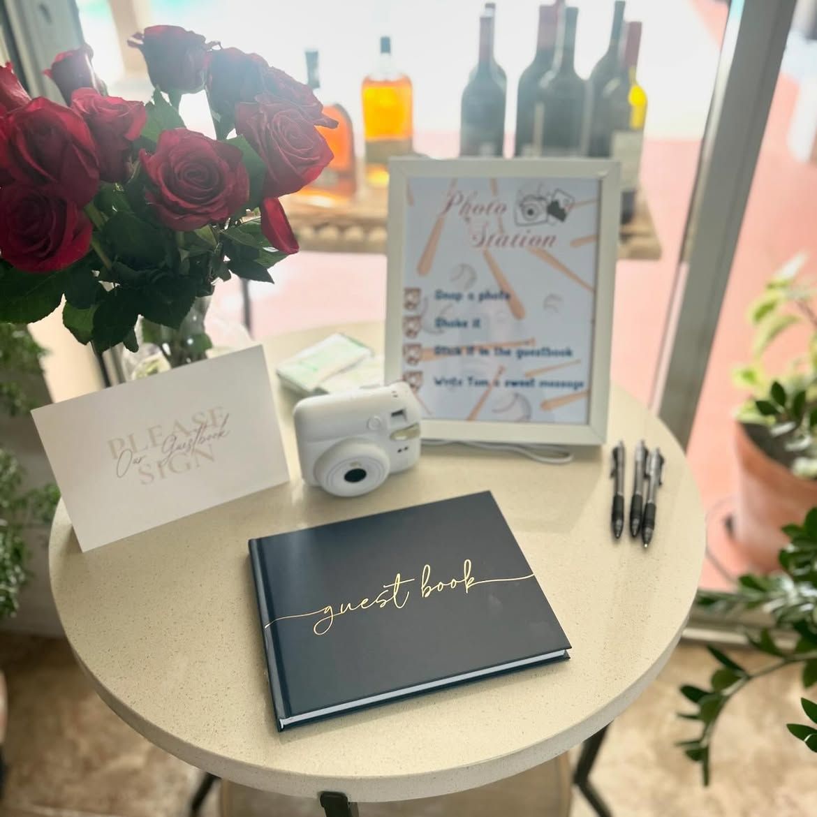 Wedding guestbook and decor table with roses, cameras, signs, and bottles on a round table.