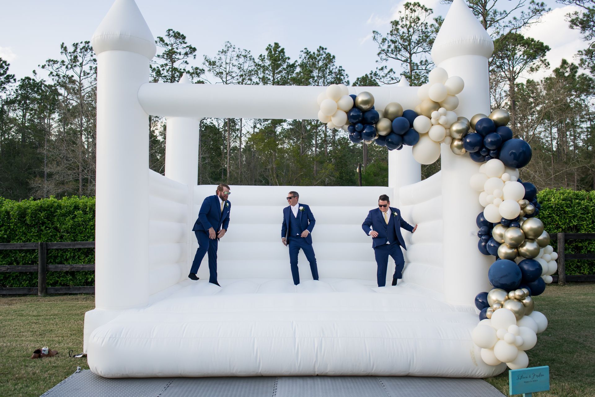 White inflatable bounce house with blue-and-gold balloons and three people in blue suits posing inside.