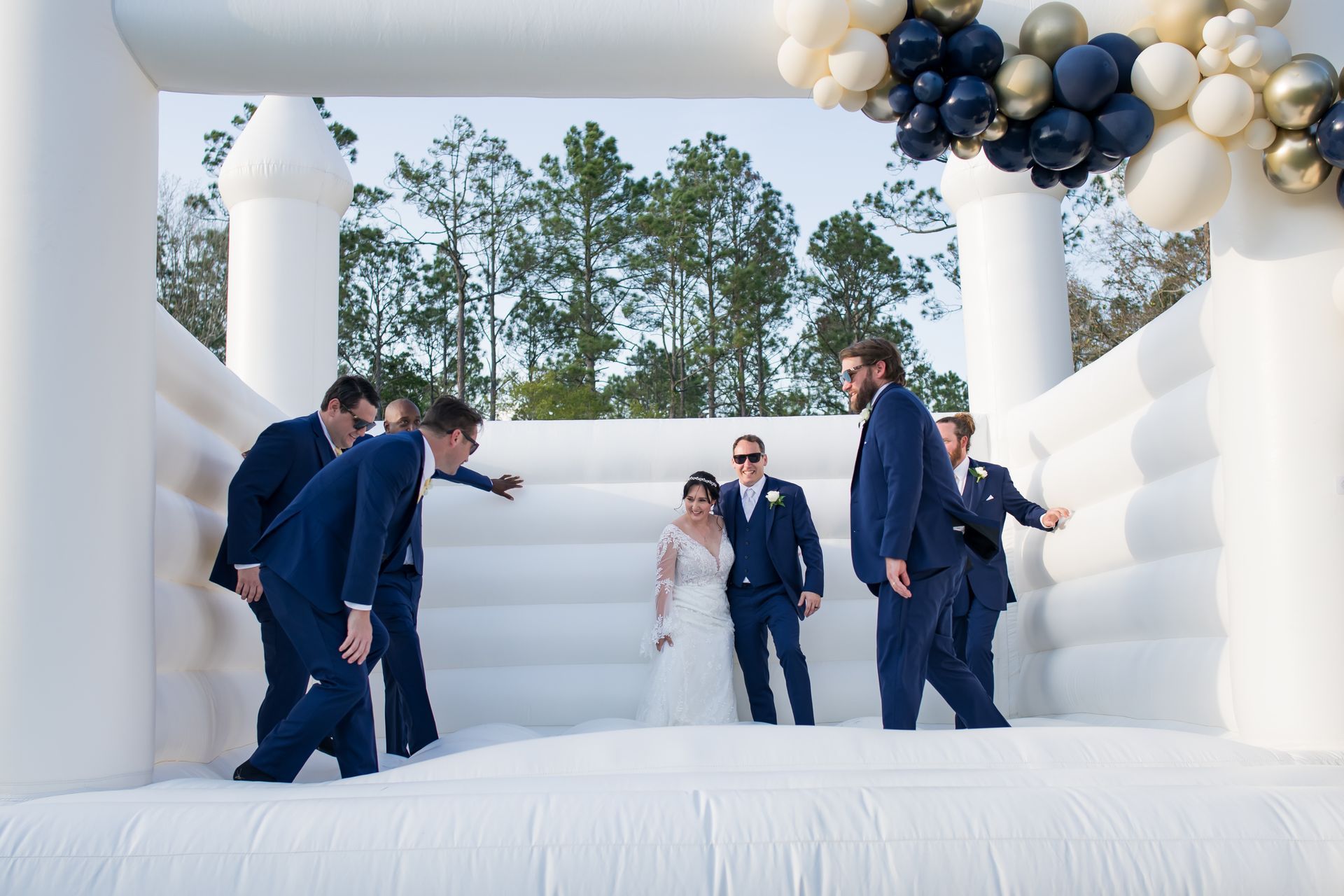 Men in navy suits gather on a white inflatable stage under black, gold, and white balloon arches outdoors