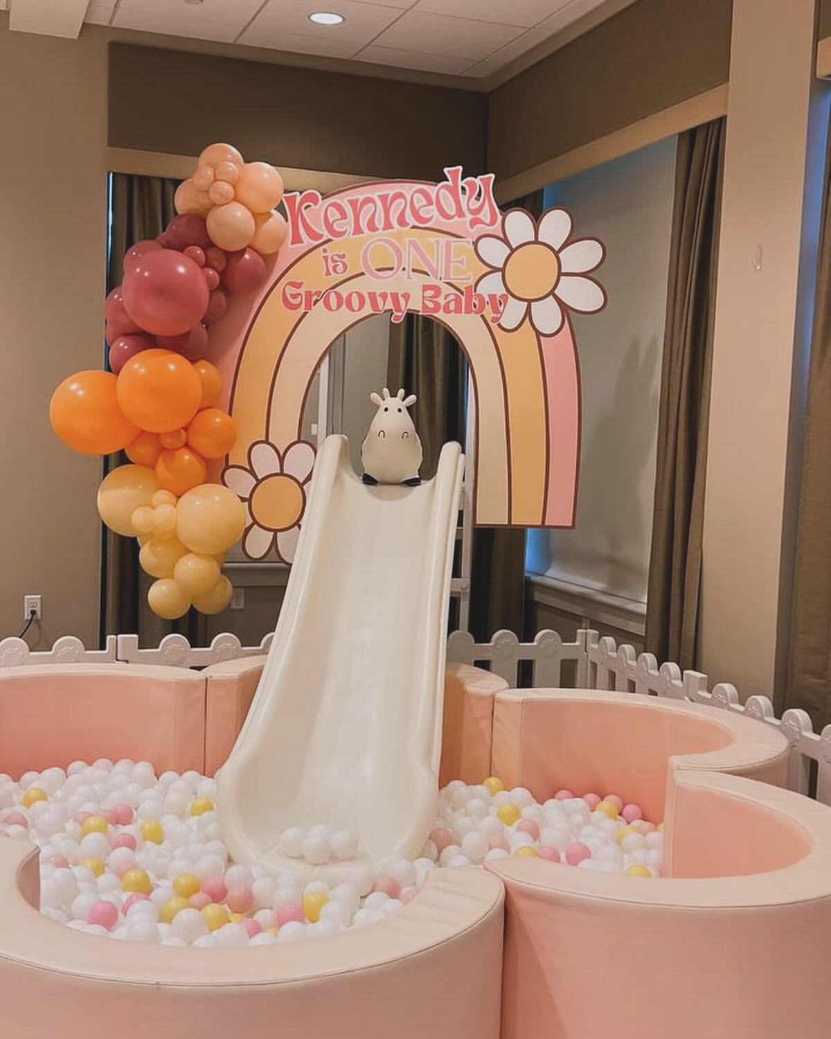 Pastel balloon-garland birth-baptism display with rainbow arch and white slide into a pink ball pit