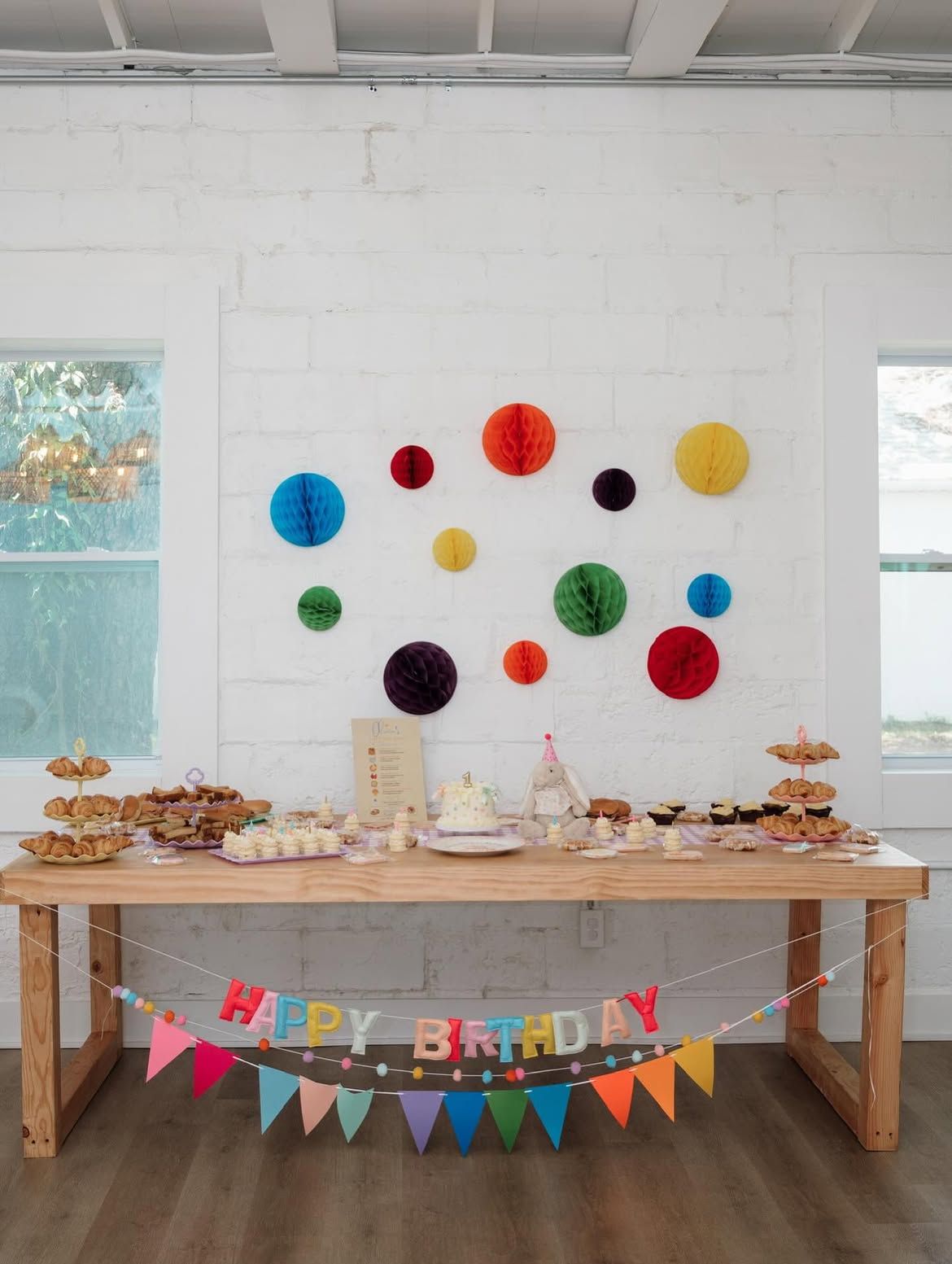 Birthday dessert table with colorful paper pom-poms and a “Happy Birthday” banner