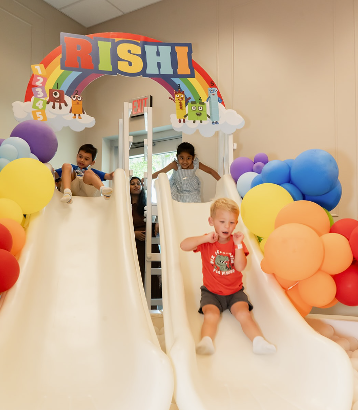 Children sliding on a birthday-themed indoor play slide with colorful balloons and a RISHI arch