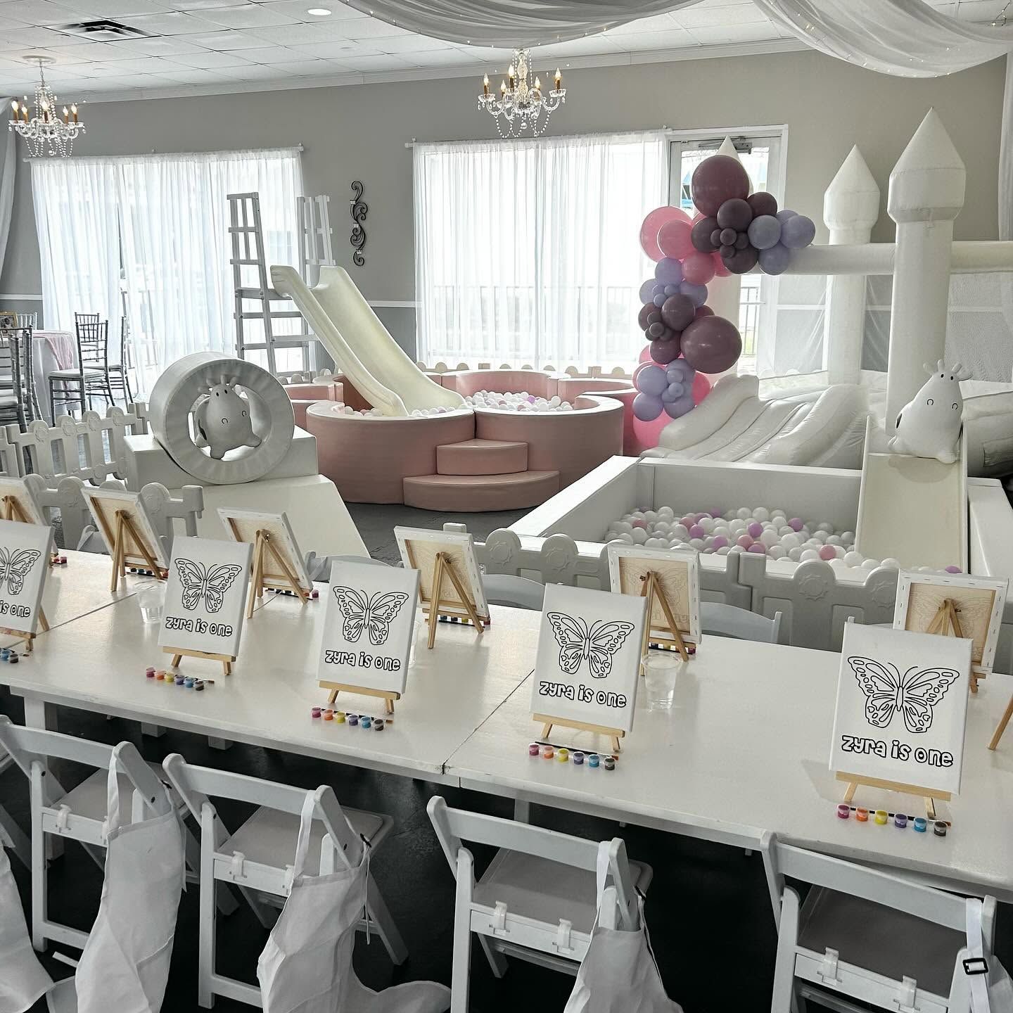 Elegant party room with long white tables, balloons, and pink-and-white inflatable play structures