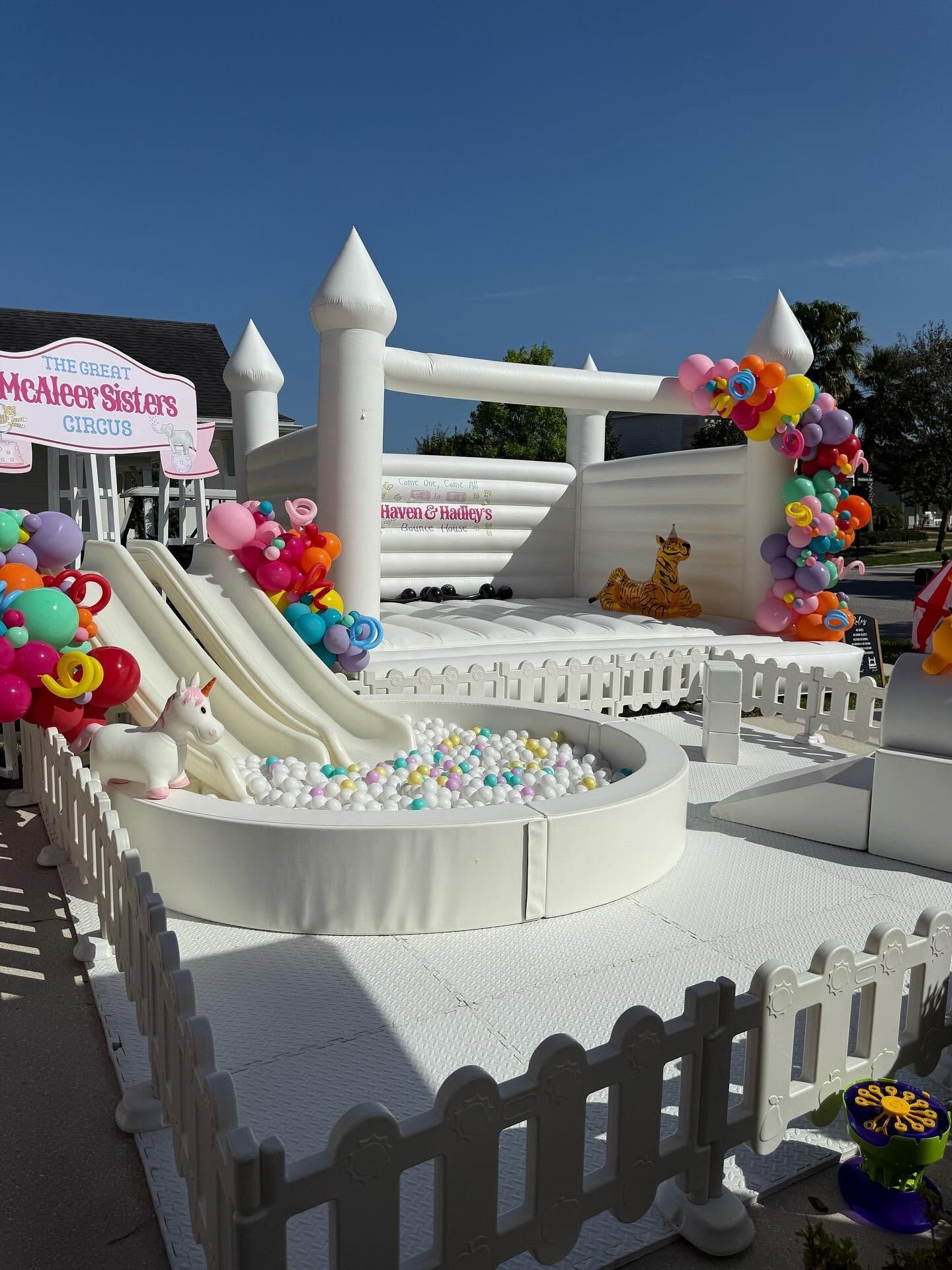 Candy-themed inflatable bounce house with colorful balloons, white picket fence, and ball pit outdoors under blue sky