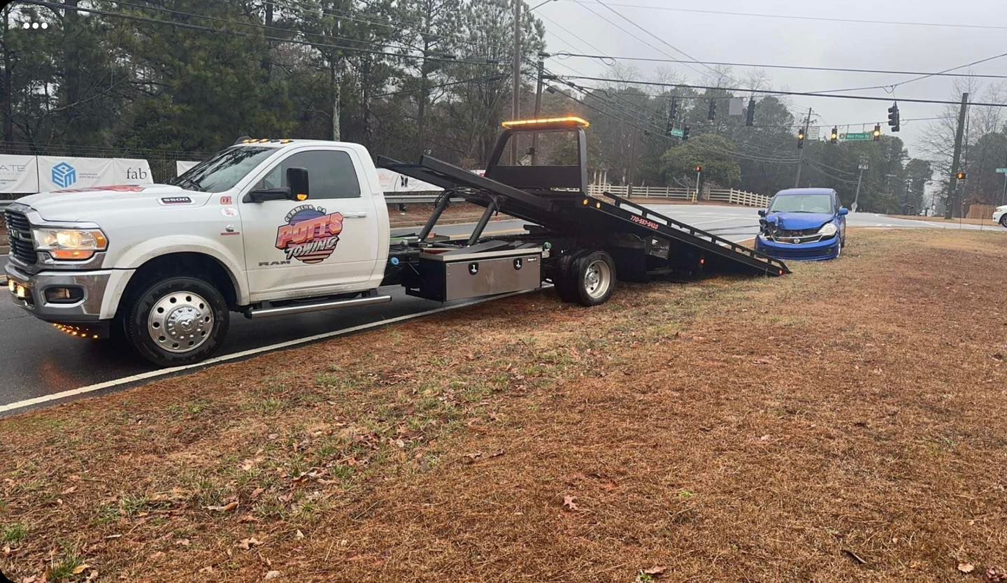 Tow truck with a flatbed on a wet roadside, carrying a car, with a blue car behind in the rain.