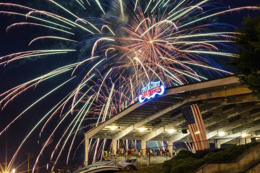 Fireworks exploding over a stadium at night, with a glowing sign and American flag on the stands