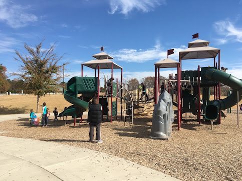 Children at a playground with green slides and climbing structures on a sandy lot under a blue sky